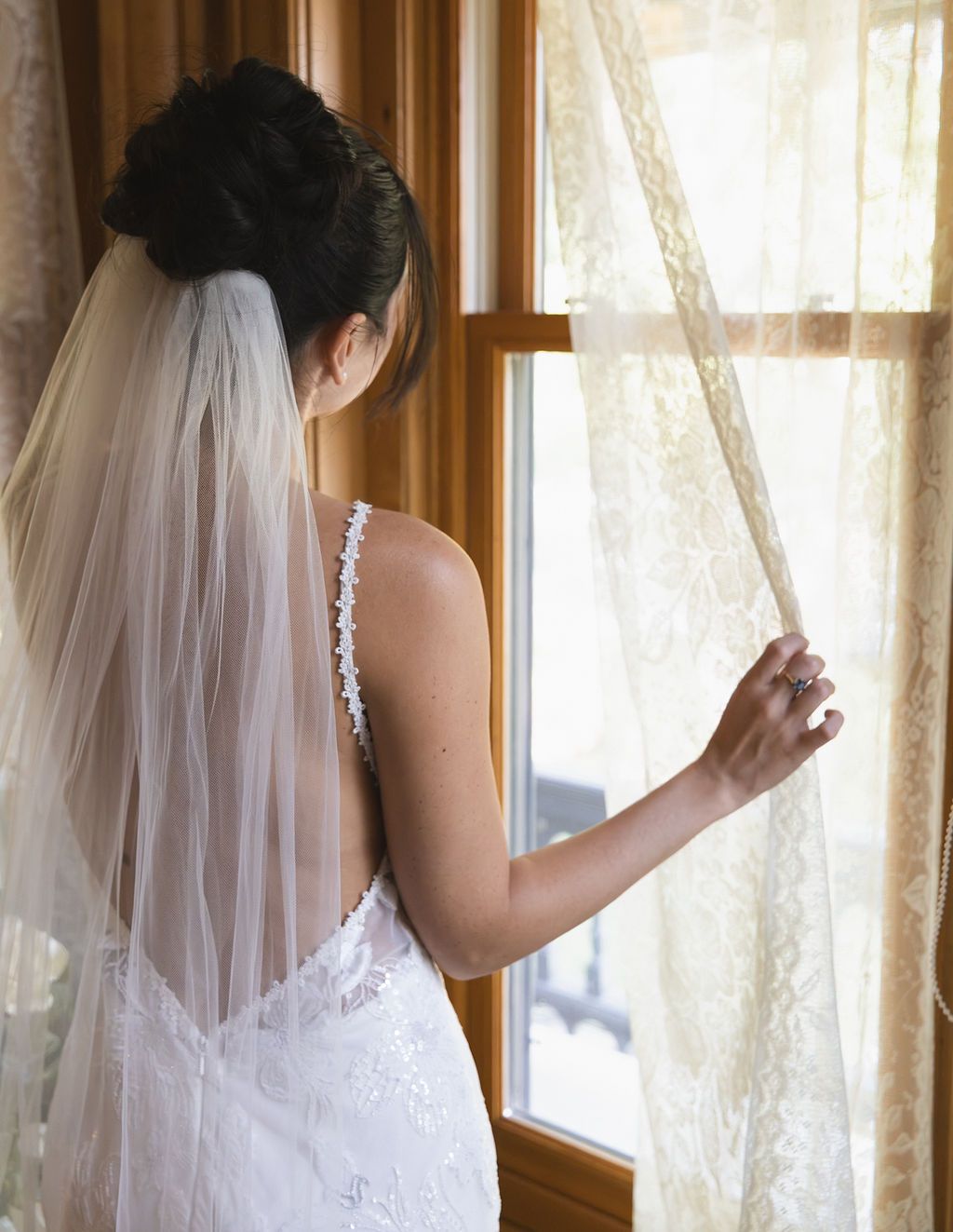 Bride in a white lace dress and veil opening a window by sheer curtains