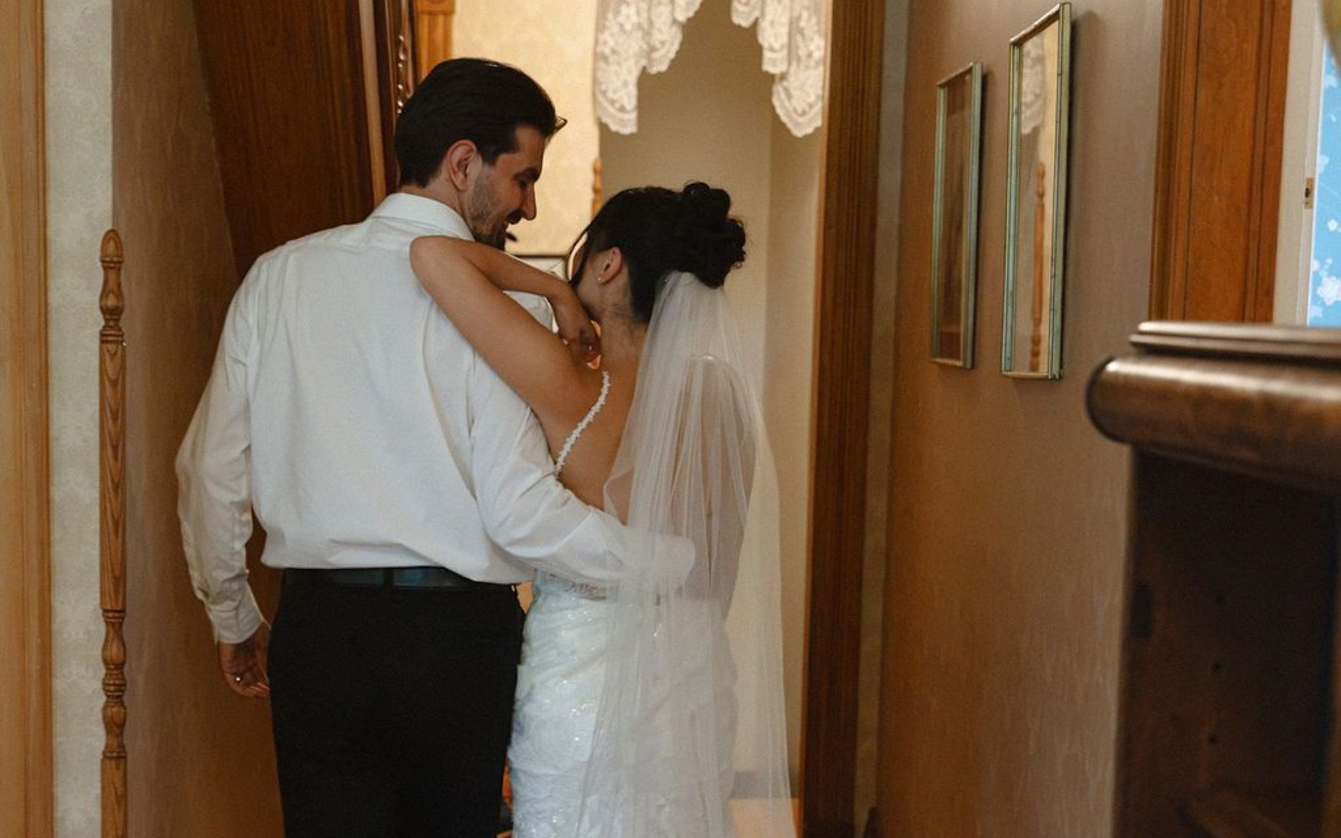 Bride and groom embrace in a hallway, dressed in wedding attire, near a lace curtain.