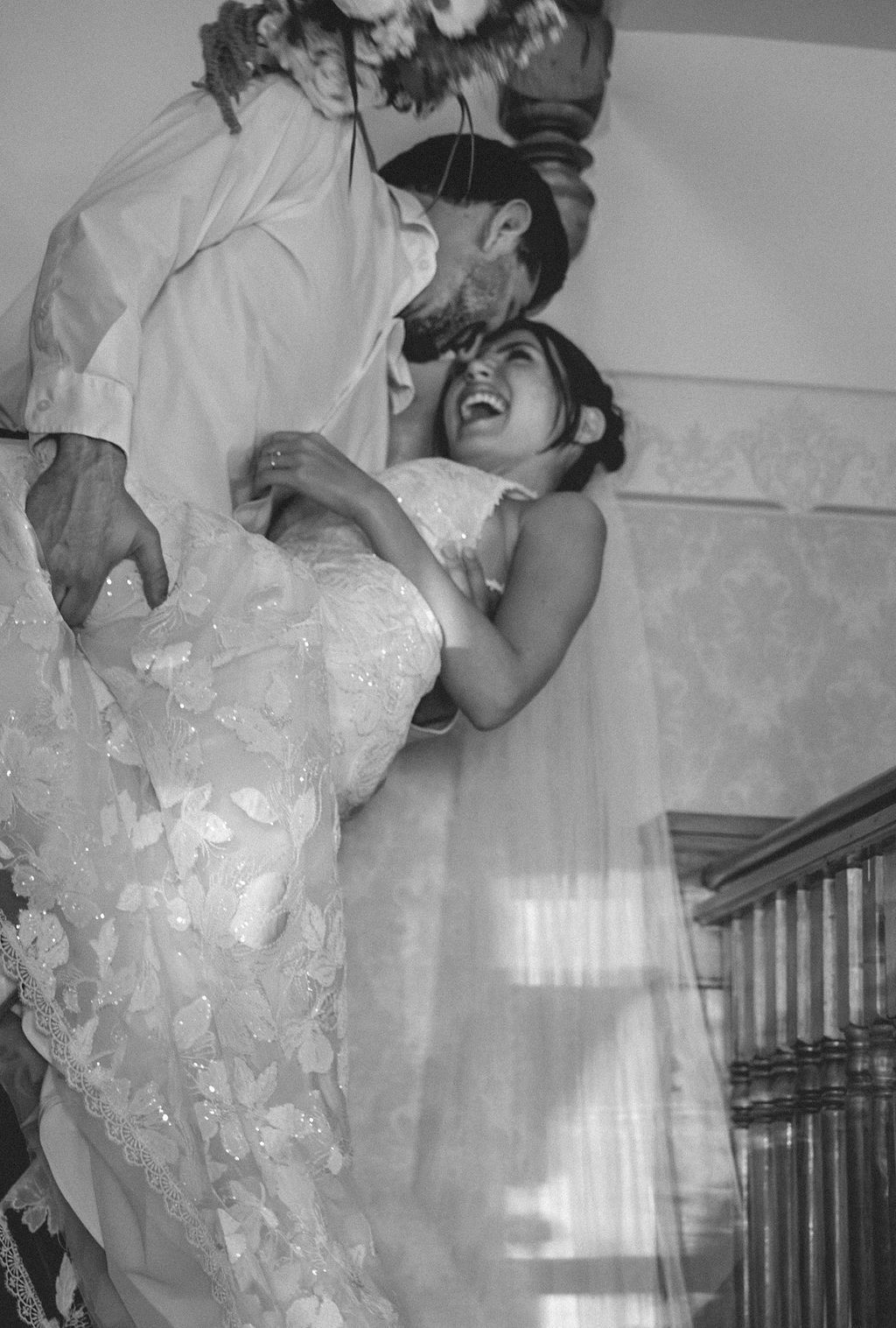 Black-and-white indoor photo of two people in formal attire, one lifting the other near a staircase