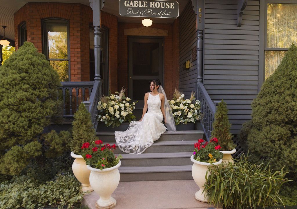 Bride in white dress sitting on steps outside Gable House, framed by flowers and greenery