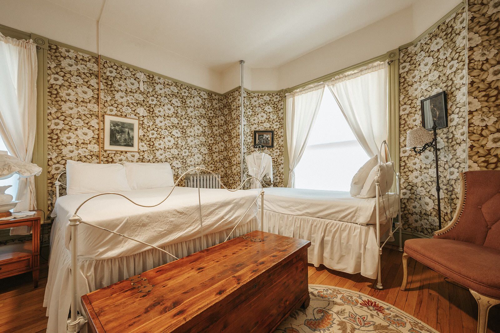 Elegant bedroom with two cream beds, floral wallpaper, a wooden chest, and curtained windows