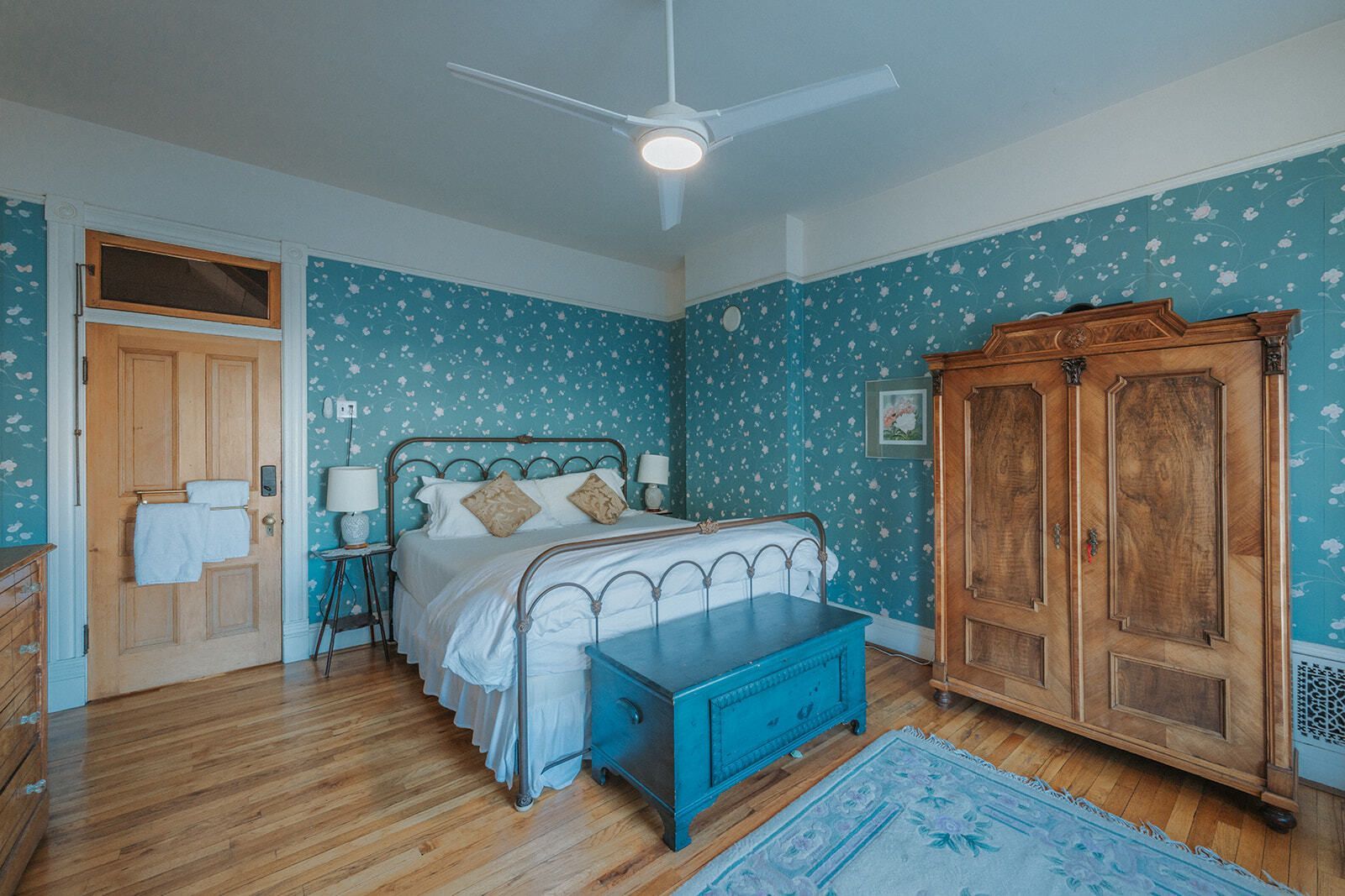 Bright bedroom with a metal bed, blue wallpaper, wooden wardrobe, and a ceiling fan.