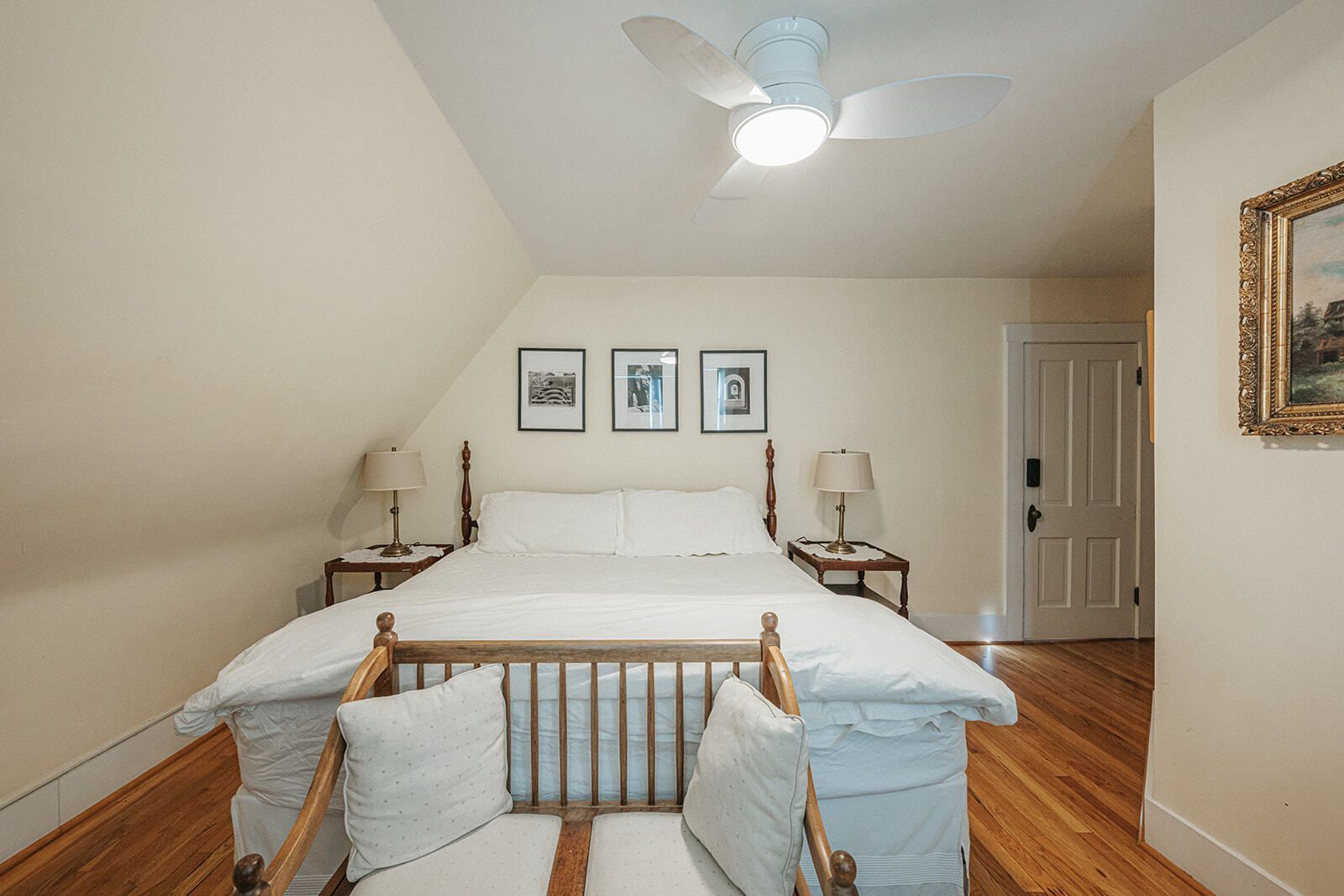 Bright bedroom with a white bed, wooden floor, ceiling fan, framed art, and a dresser by the door.