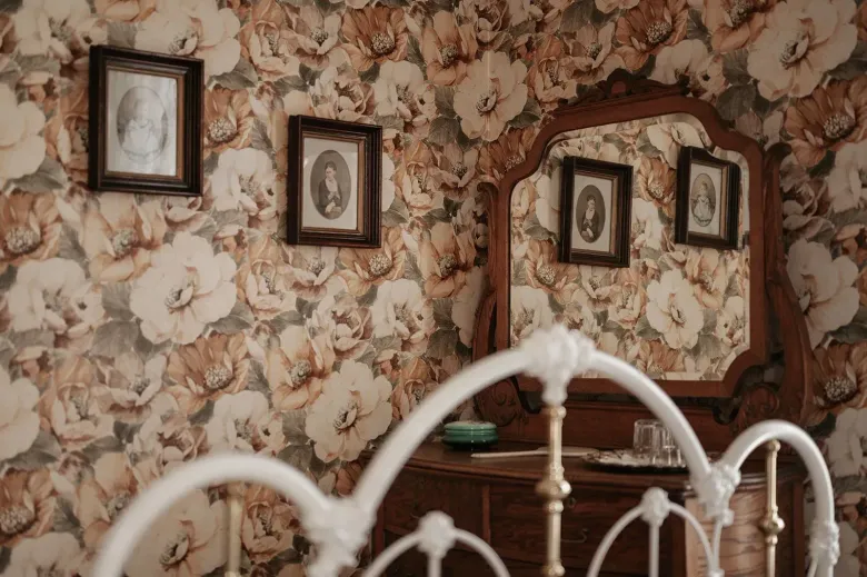 Floral wallpapered bedroom with a white metal bed frame and framed portraits on the wall