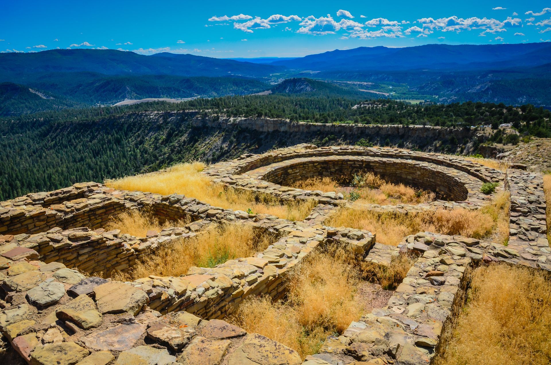 Ancient stone ruins on a hillside overlooking a forested valley and distant mountains under a blue sky
