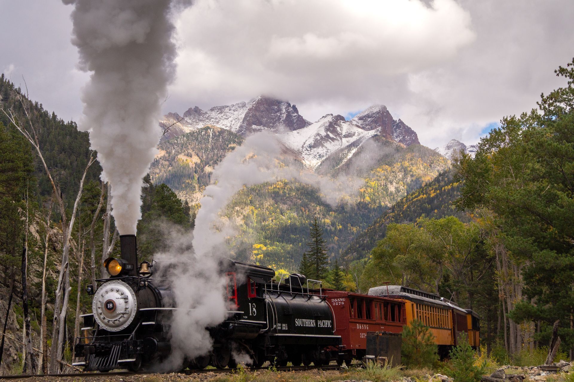 Steam locomotive pulling red-orange cars through a mountain forest with snowy peaks in the background