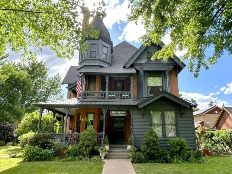 Victorian house with gray siding, wraparound porch, and lush green yard under trees.