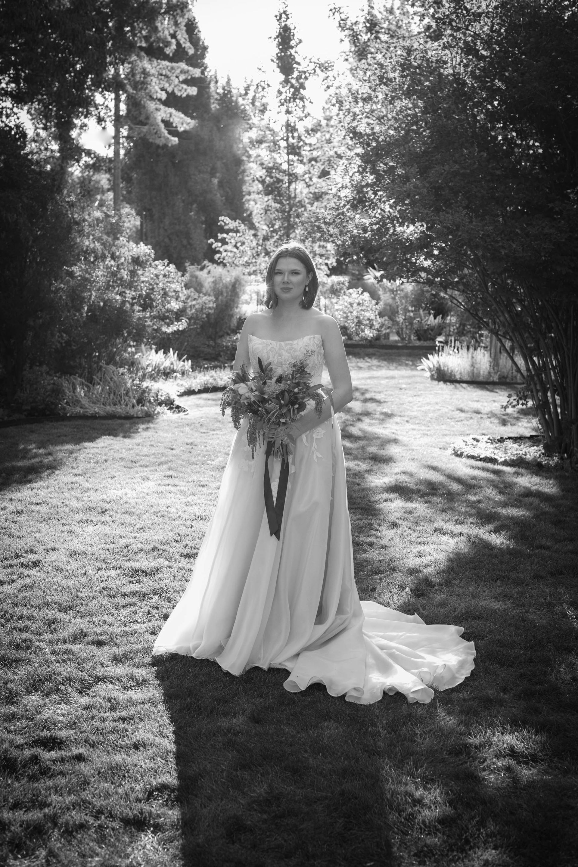 Bride in a long white gown holding a bouquet, standing in a sunlit garden path.