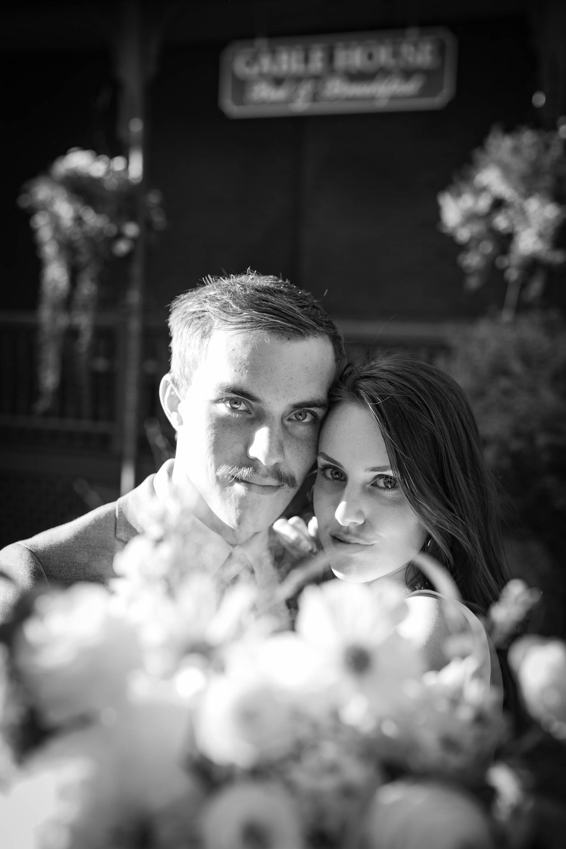 Couple posing closely outdoors with blurred flowers in the foreground, black-and-white portrait