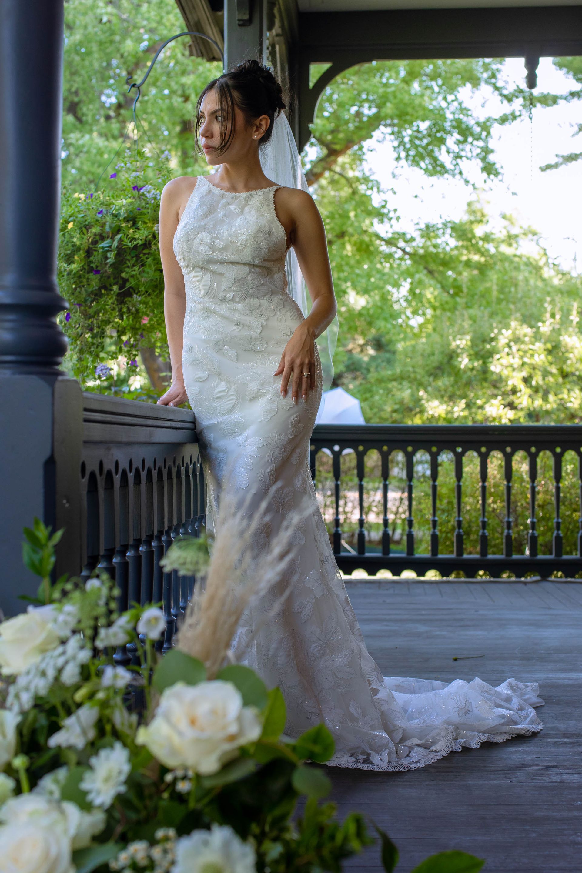 Bride in a white lace gown standing on a porch with flowers in the foreground.