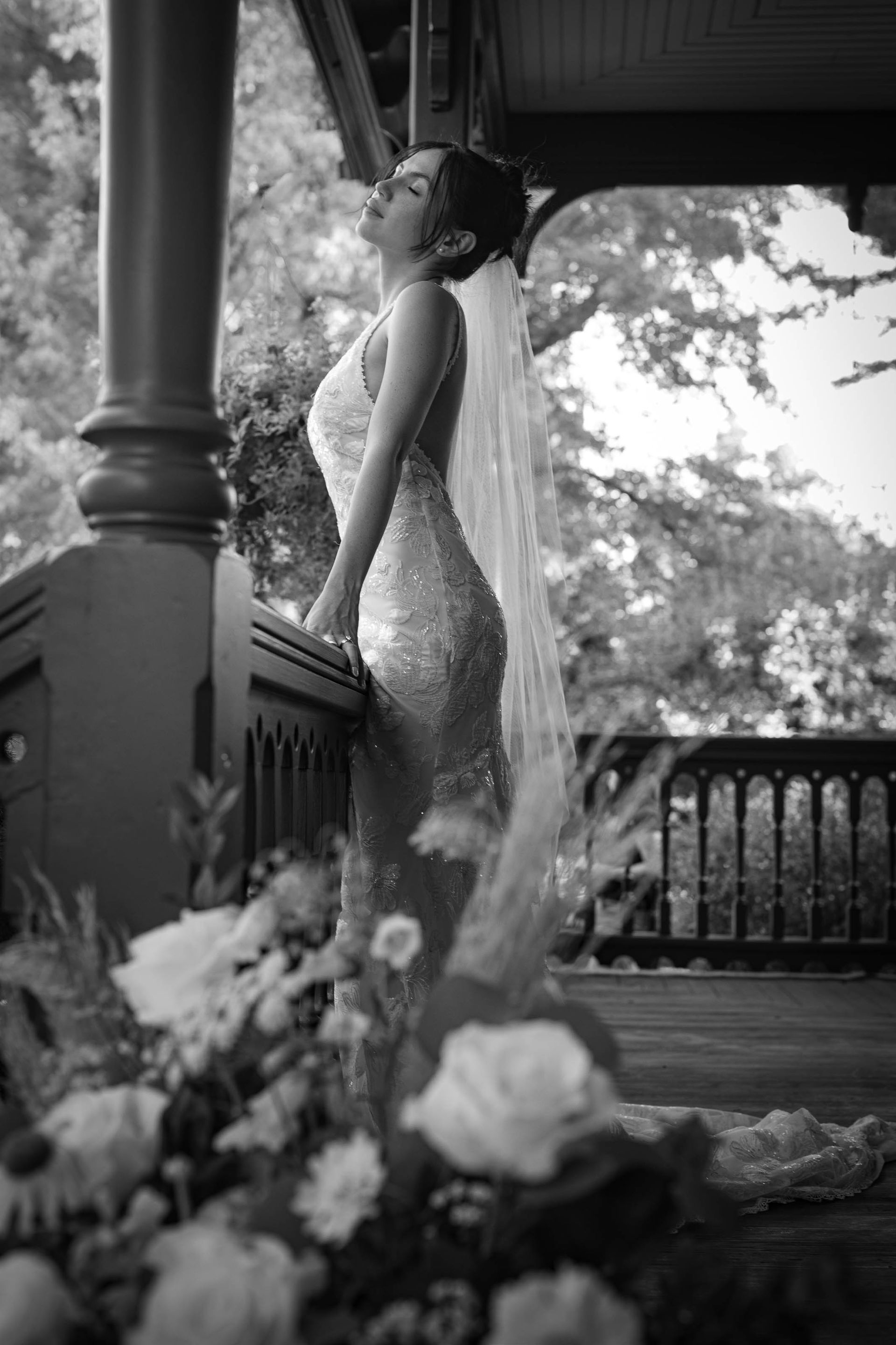 Woman in a shimmering gown standing on a porch beside flowers, looking upward.