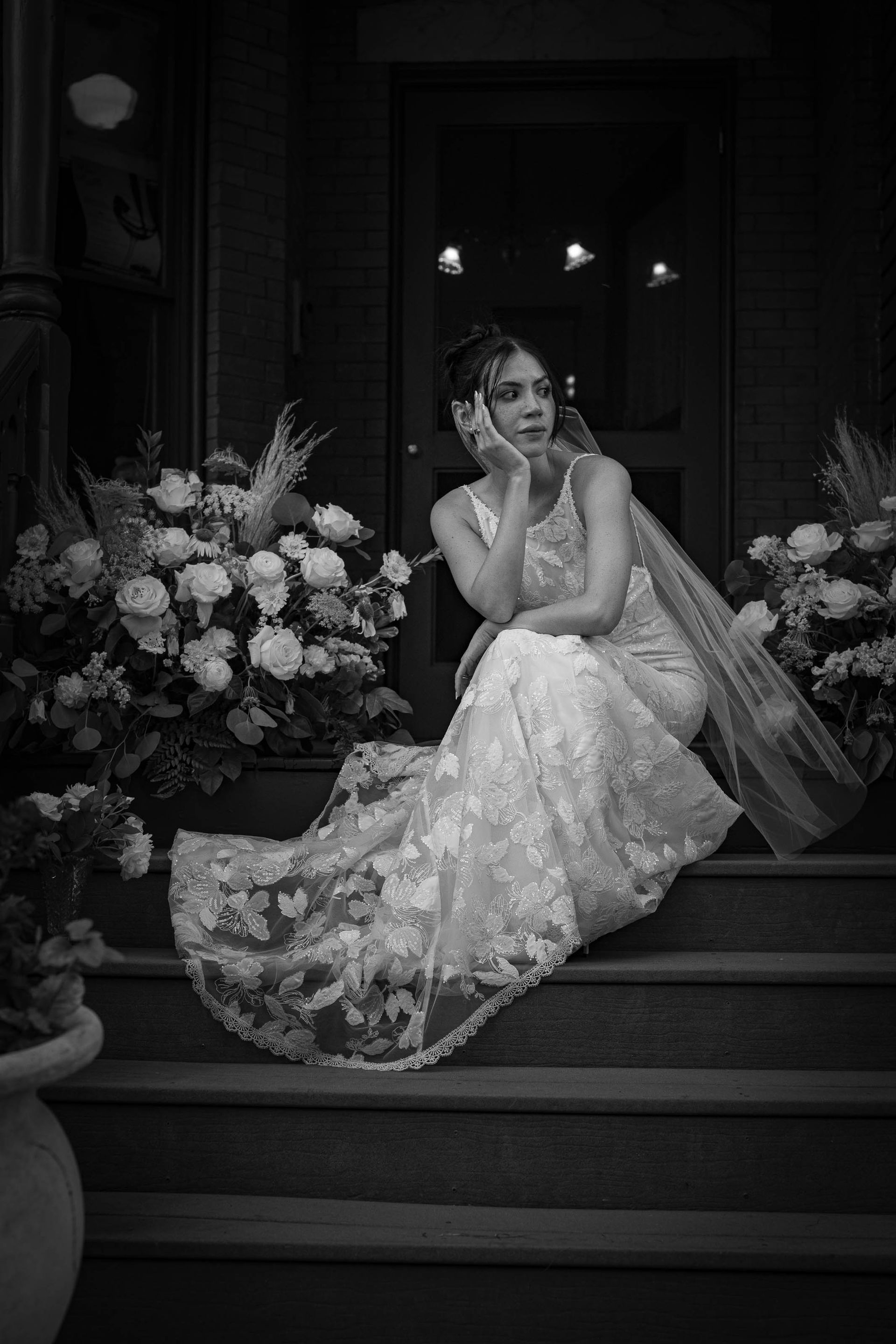 Bride in a lace gown sitting on steps beside floral arrangements, posing in a doorway