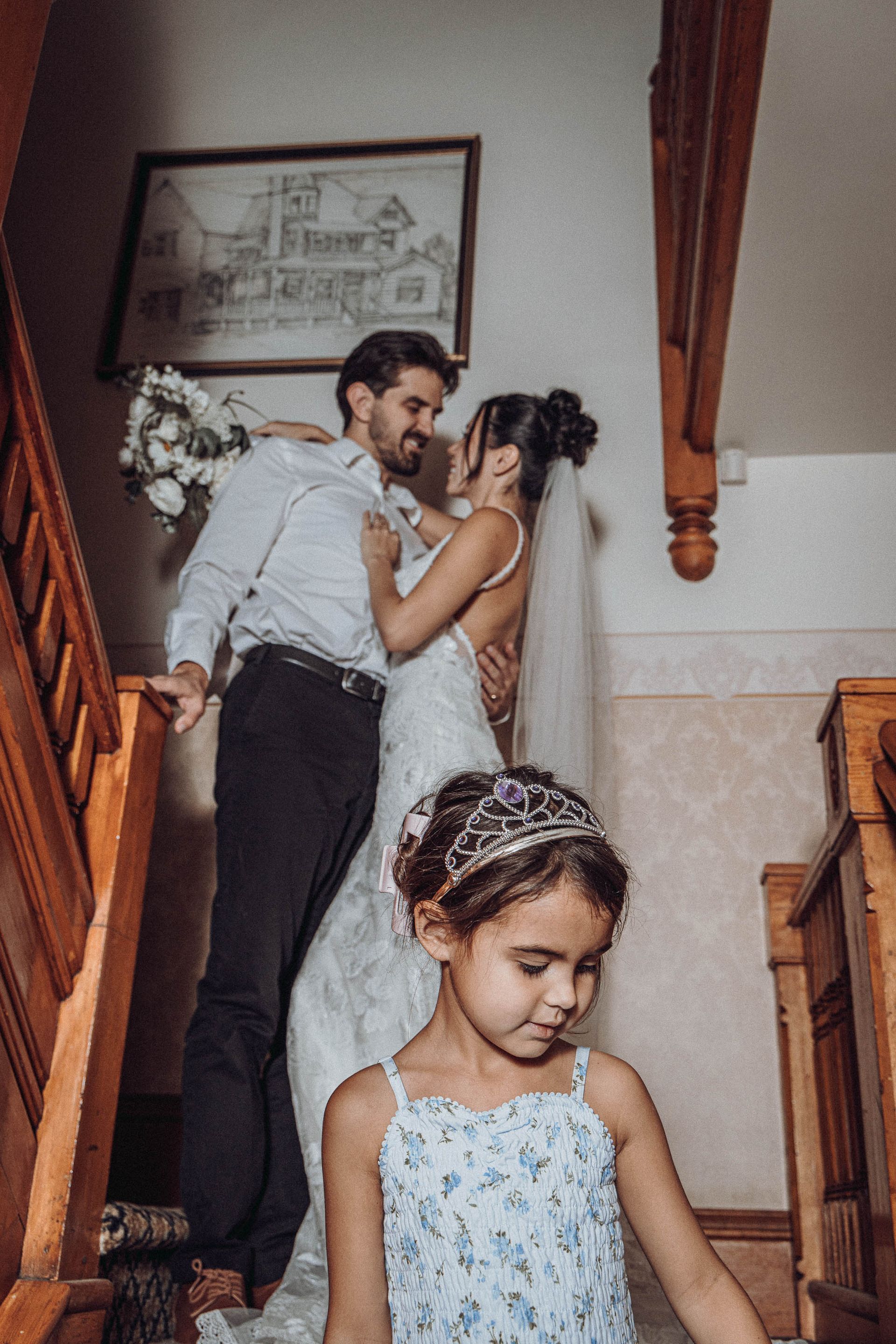 Bride and groom kiss on a staircase while a young flower girl stands in front.