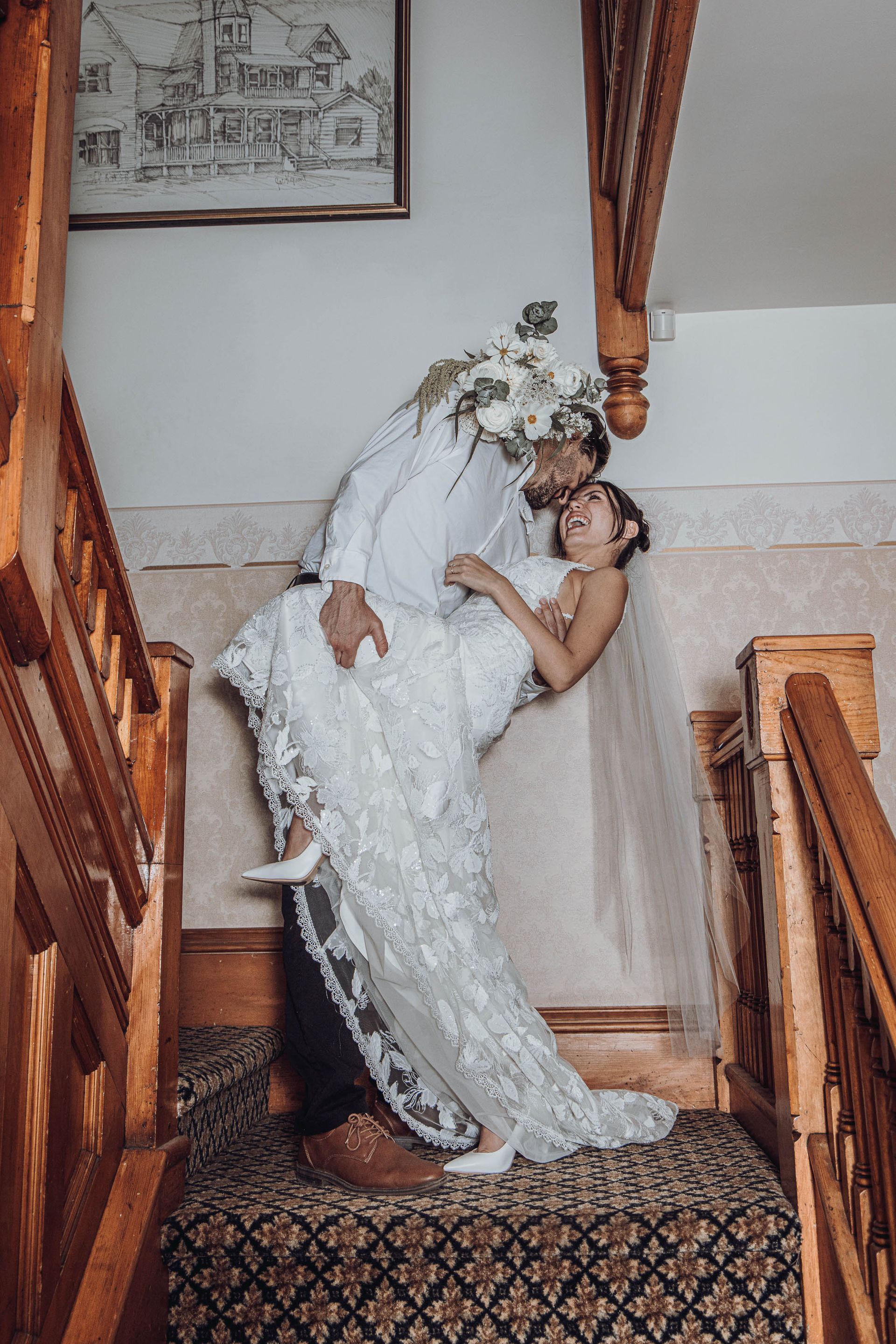 Bride in white gown being lifted on a staircase by another person, both smiling