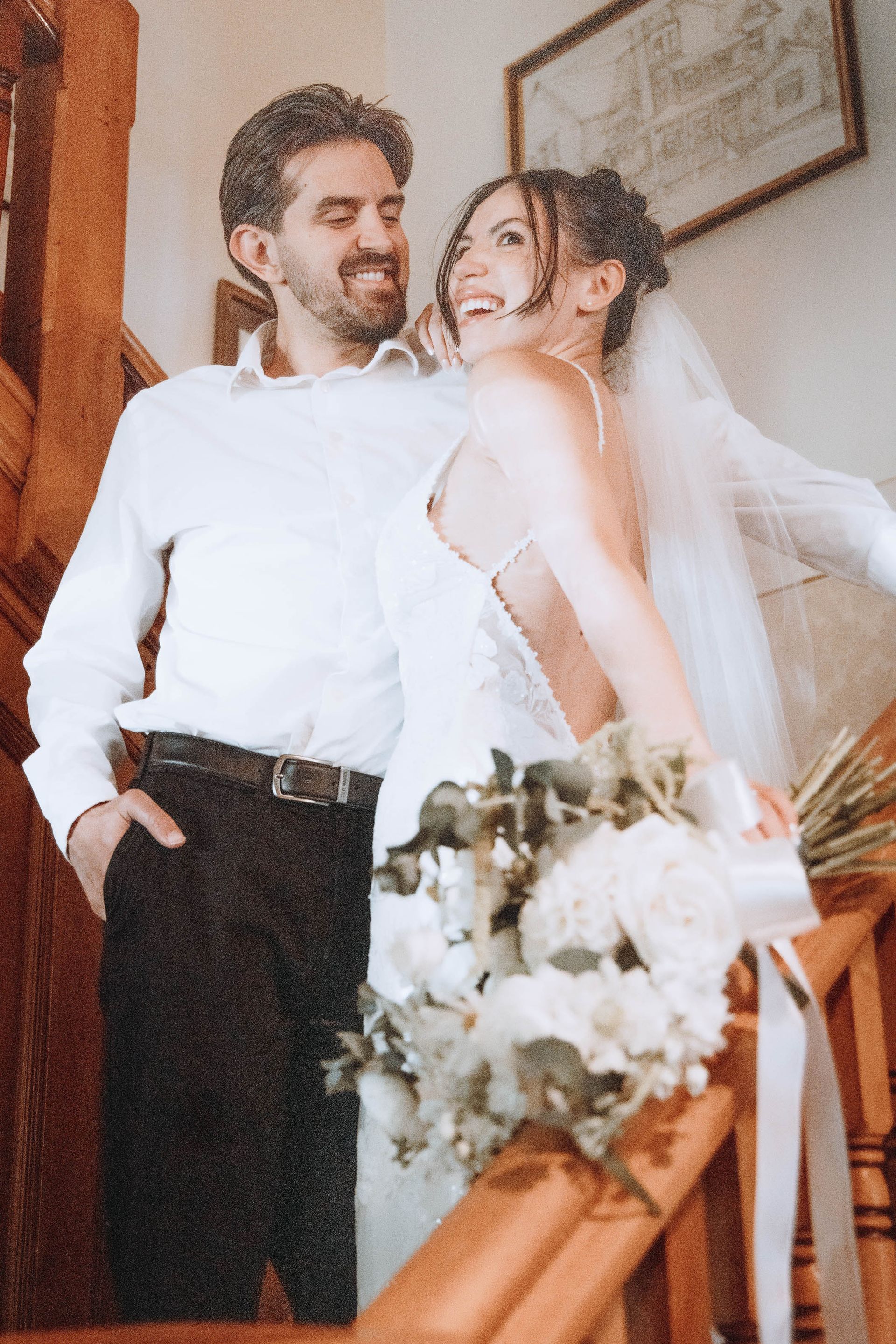 Smiling couple in wedding attire posing indoors, with bouquet and veil visible