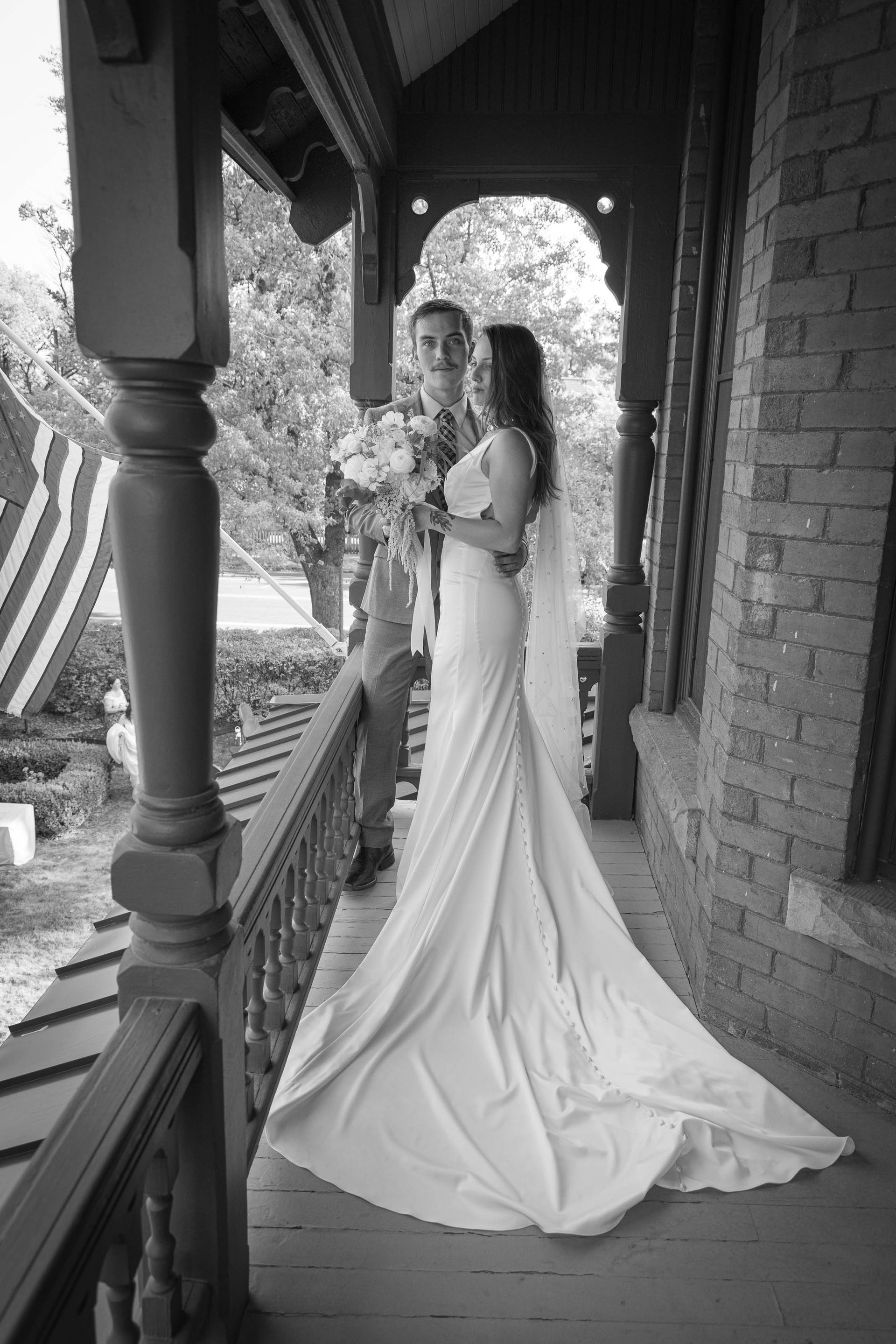 Bride in a long white gown standing on a porch beside a brick wall, holding a bouquet.