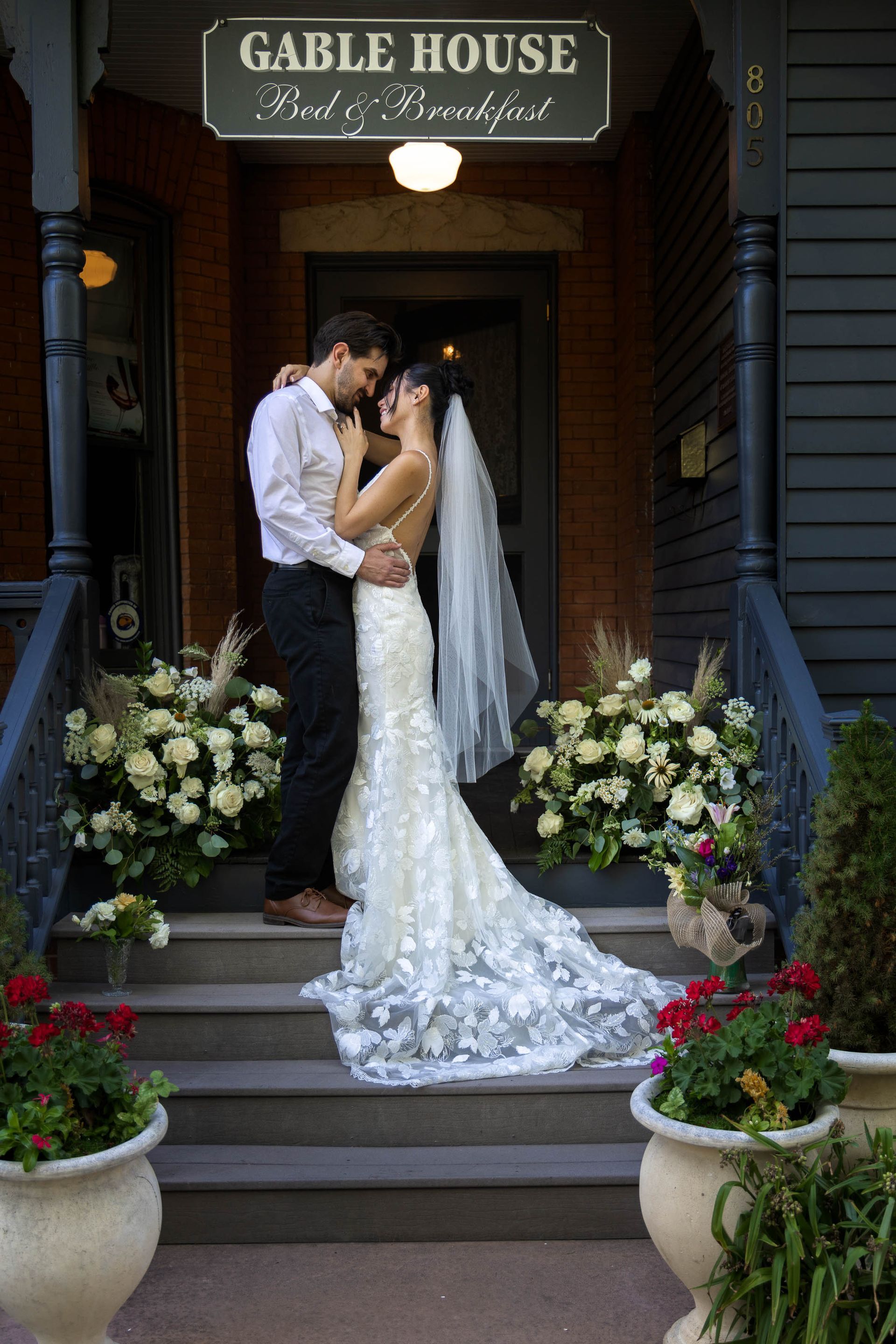 Wedding couple kissing on the steps outside Gable House, with white flowers and the bride’s long veil trailing behind