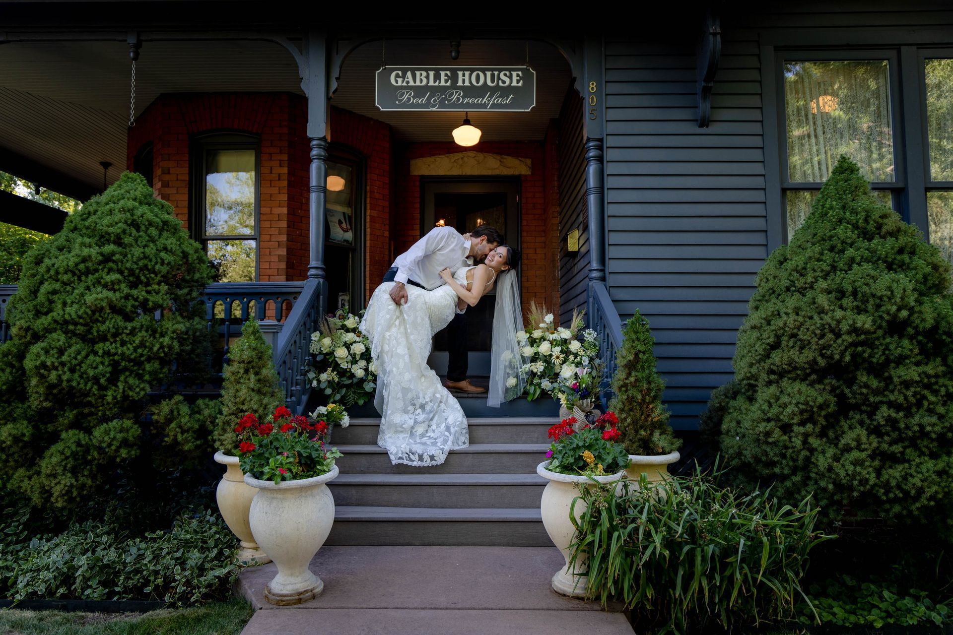 Bride kissing partner on steps outside Gable House, framed by flowers and greenery