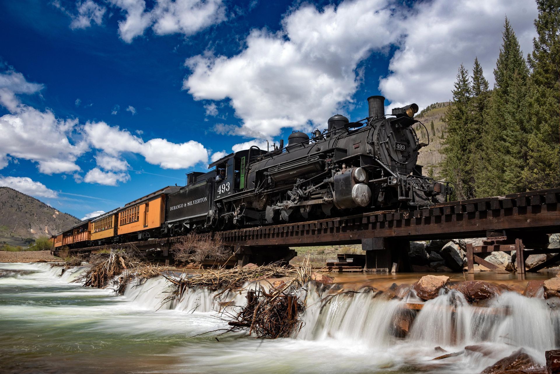 Steam locomotive crossing a bridge over a rushing river under a cloudy blue sky