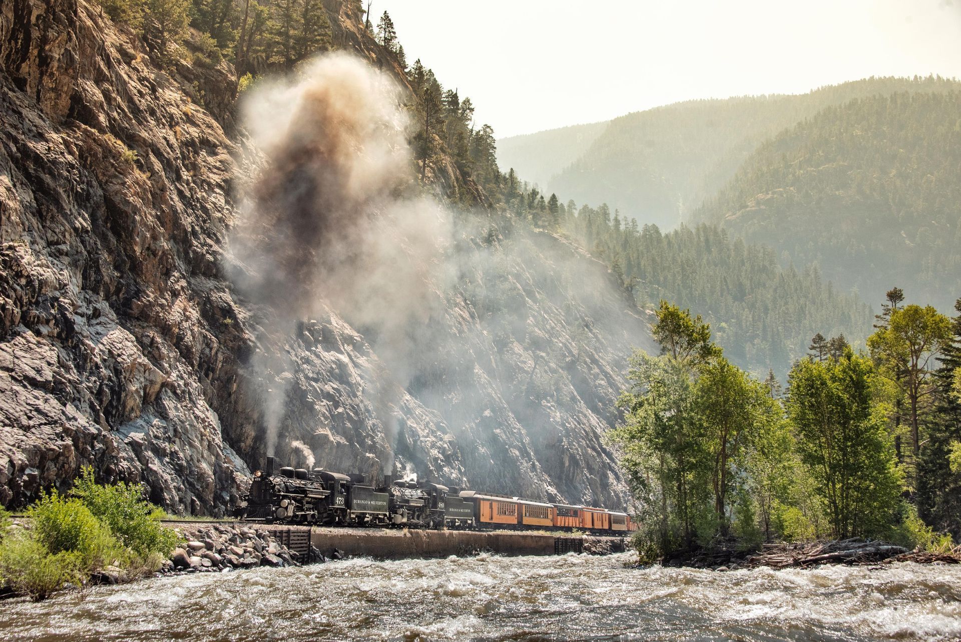 Steam train crossing a rocky mountain gorge, with smoke rising along a river valley.