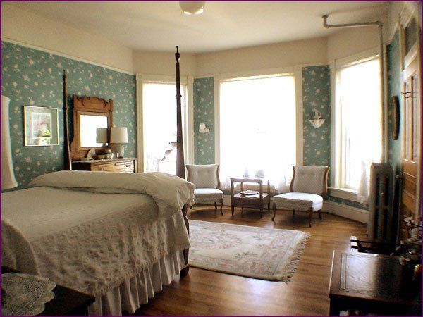Spacious bedroom with a bed, patterned wallpaper, large windows, armchairs, and a rug on hardwood floors