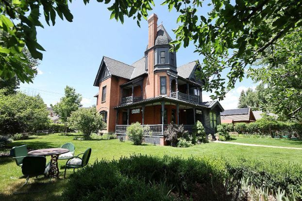 Victorian house with wraparound porch and tower, surrounded by a green lawn and trees.