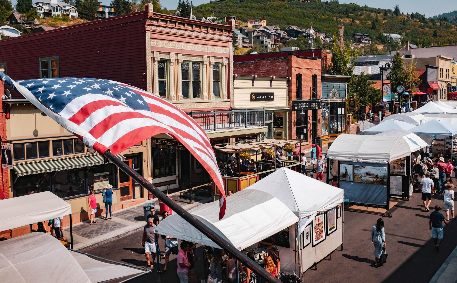 Large American flag over an outdoor street market with white tents and historic storefronts under sunny skies