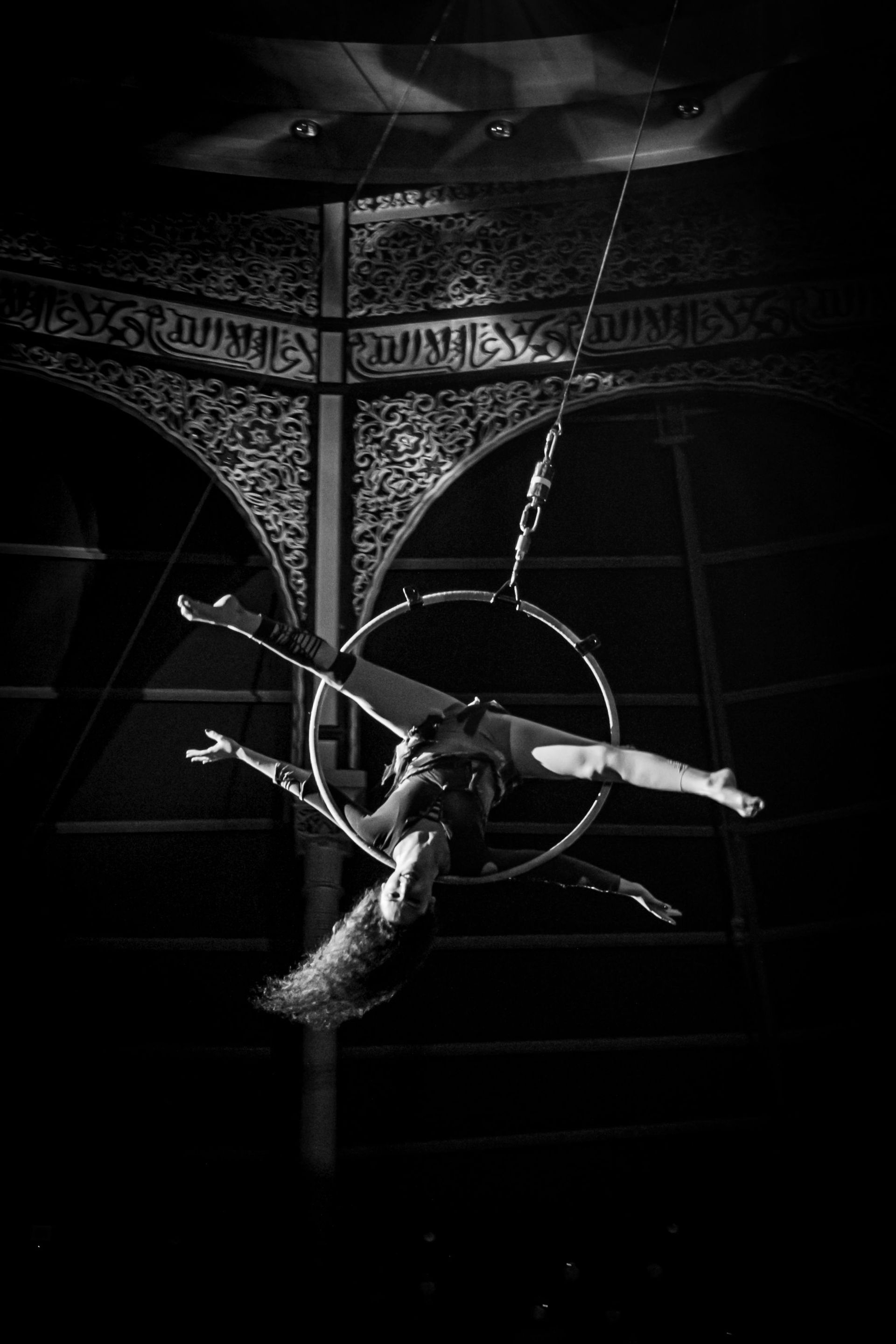 Suspended aerial performer in a hoop under ornate ceiling, lit dramatically in black and white