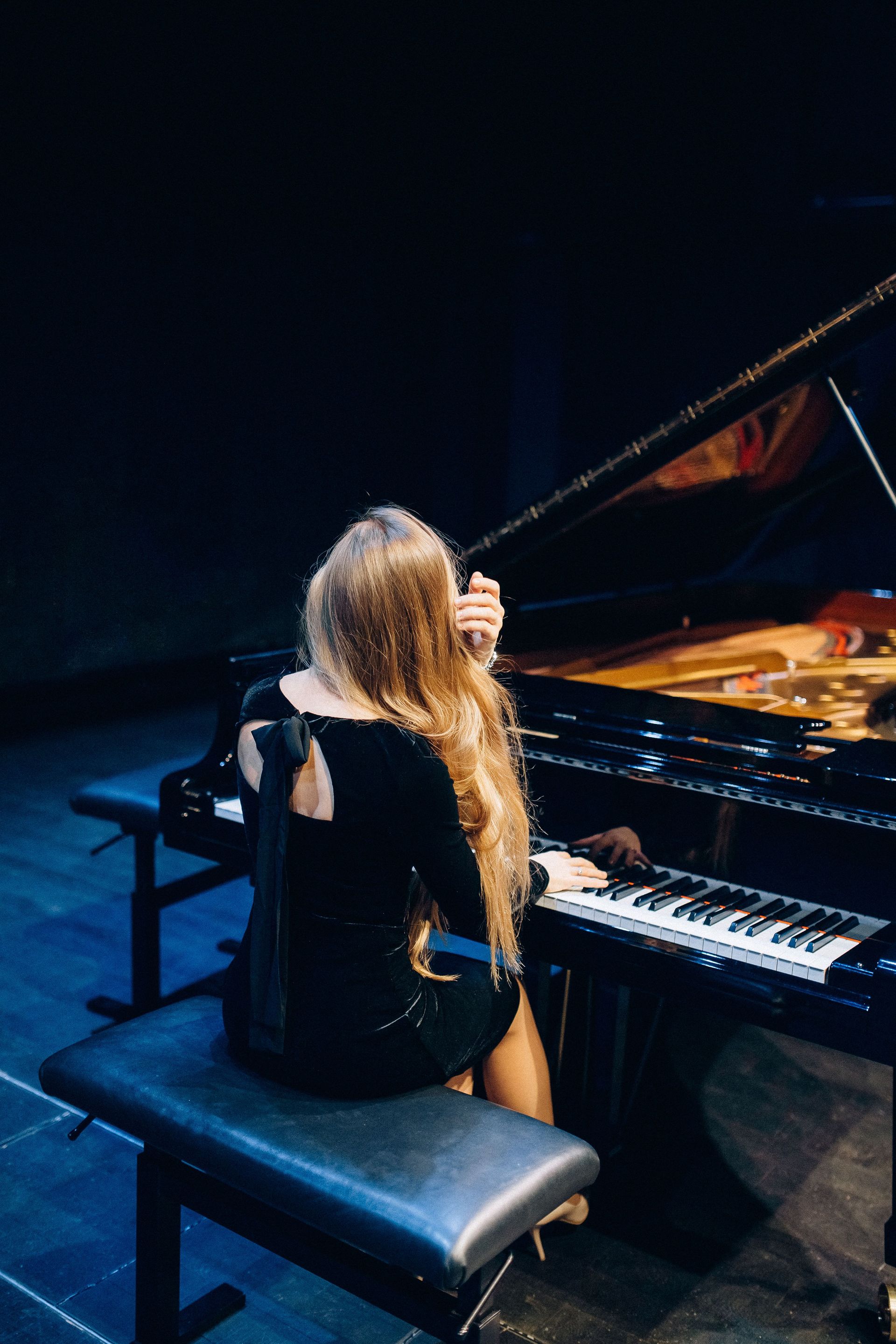 Person seated at a grand piano on a dark stage, viewed from behind