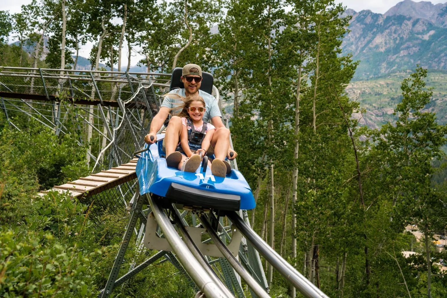 Two people ride a blue alpine coaster through a forested mountain setting.