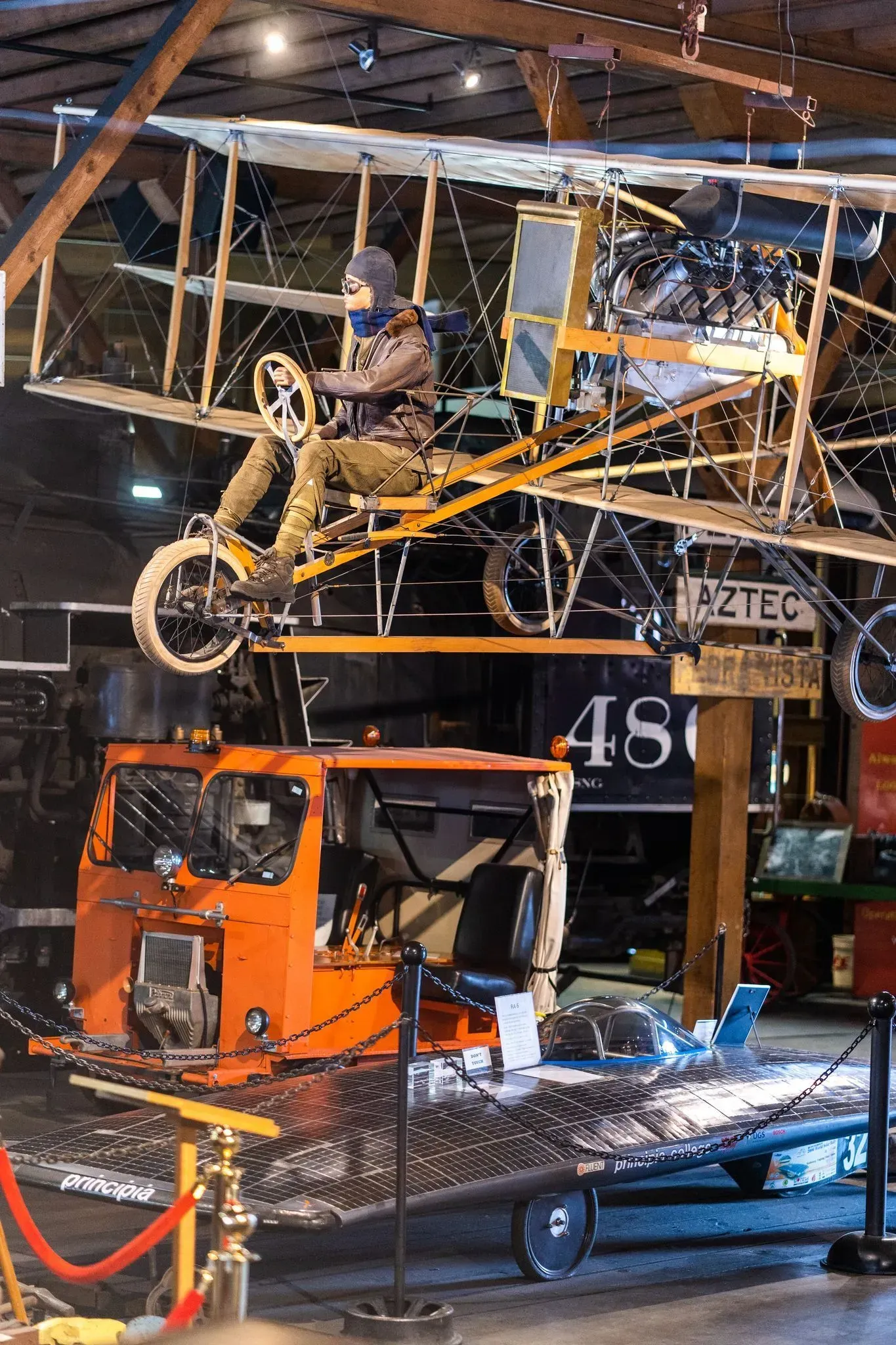 Orange industrial machine in a workshop, with a worker seated on a raised metal structure overhead.