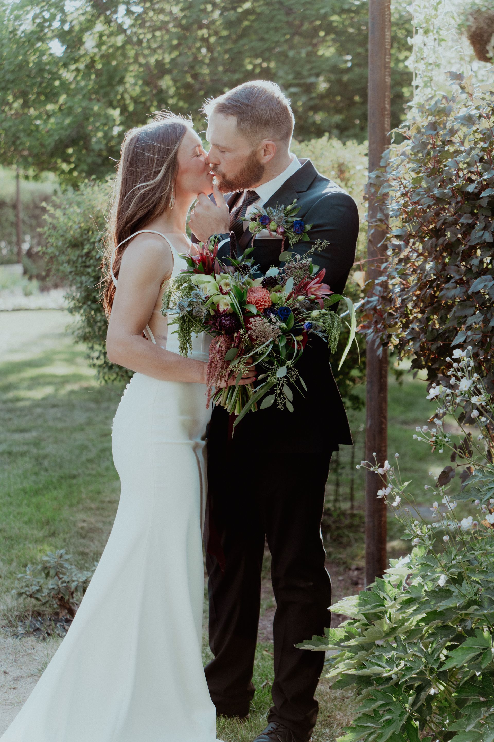 Couple in wedding attire kissing outdoors, the bride holding a bouquet of flowers.