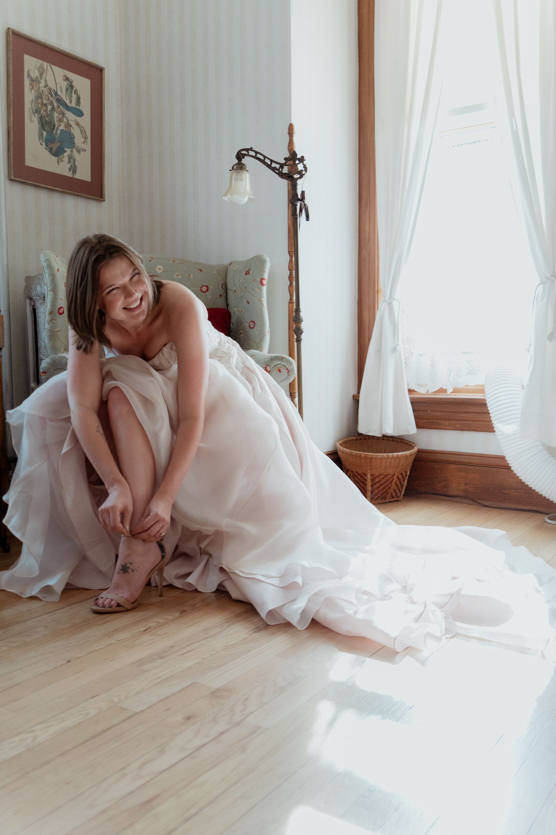 Woman in a pale pink dress sitting on a sunlit bedroom floor by a window.