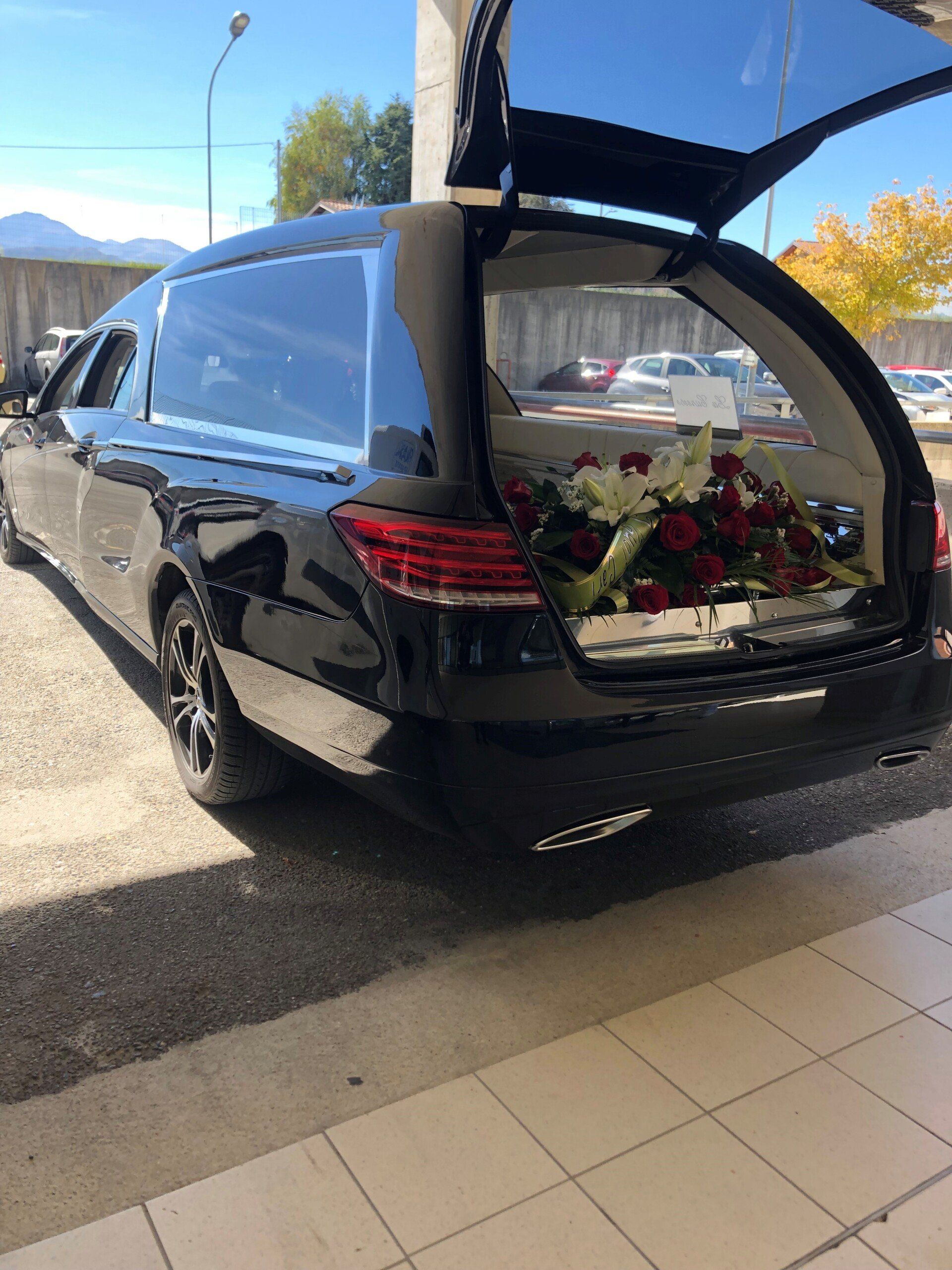 Carro funebre nero con il bagagliaio aperto, che rivela i fiori. All'aperto, giornata di sole.