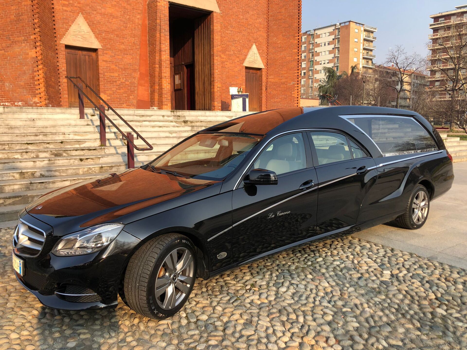 Carro funebre nero parcheggiato sul selciato di fronte a una chiesa in mattoni.