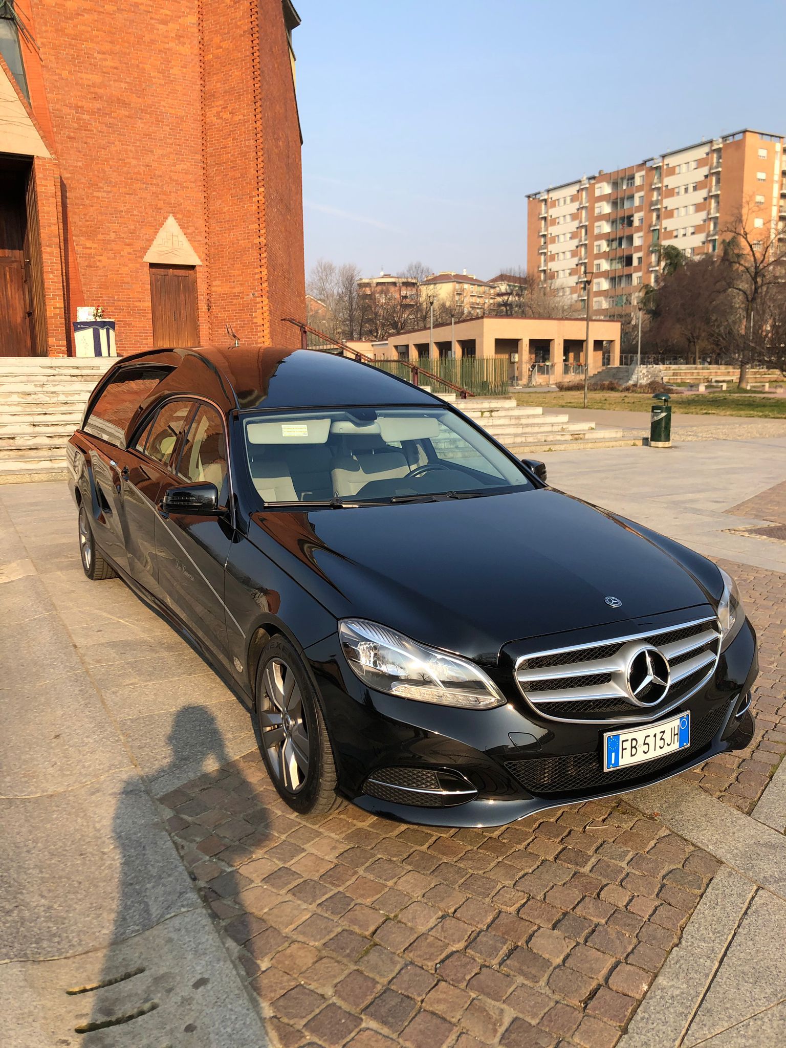 Carro funebre nero parcheggiato su un'area acciottolata vicino a un edificio in mattoni e altre strutture; giornata di sole.