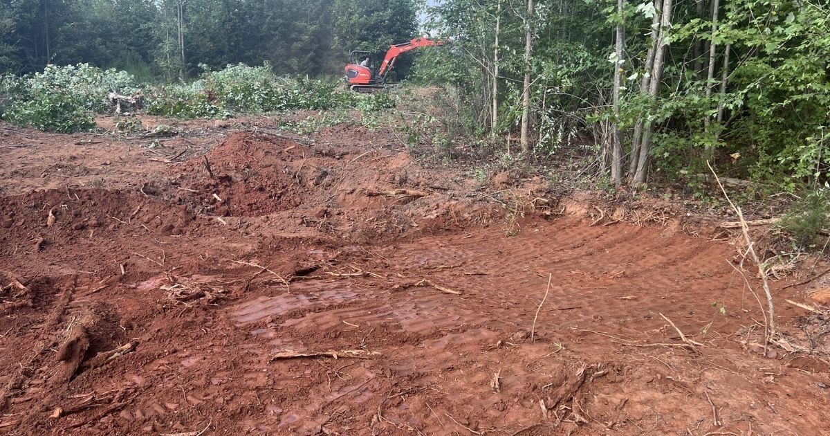Cleared red dirt lot beside woods, with a red excavator in the distance.
