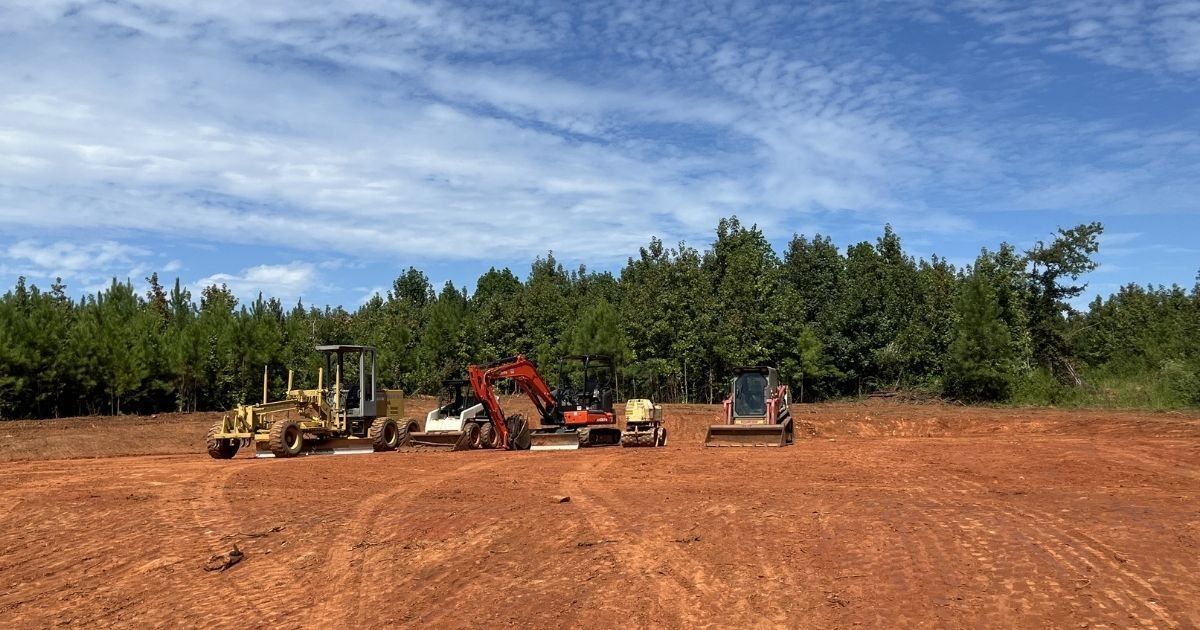 Bulldozers and a skid steer on a cleared red dirt lot beside a tree line under a blue sky
