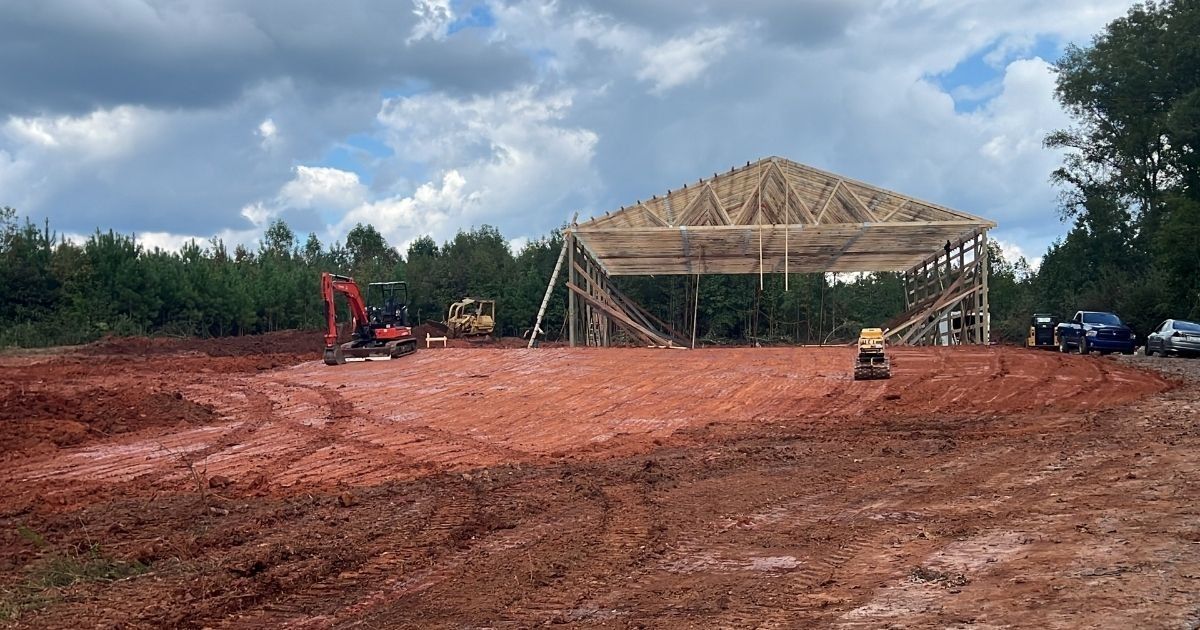Construction site with red dirt, partially built wooden pavilion, and excavator under cloudy sky