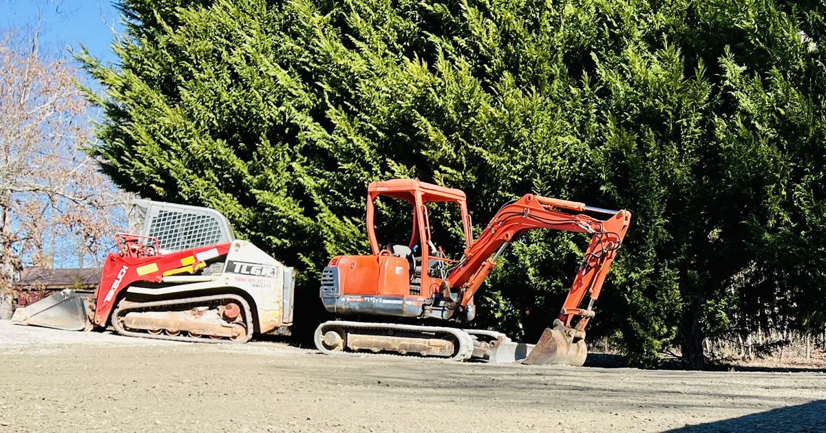 Red and orange excavators parked on dirt in front of dense green trees.