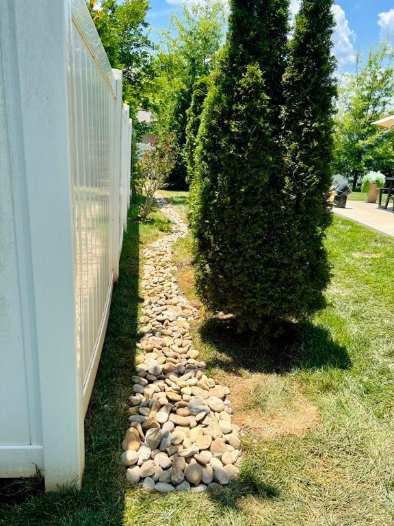Narrow rock-lined garden bed beside a white fence, with tall evergreen shrubs in a sunny yard.