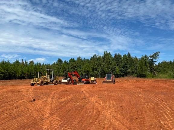 Construction equipment on a red dirt clearing under a blue sky, with trees in the background