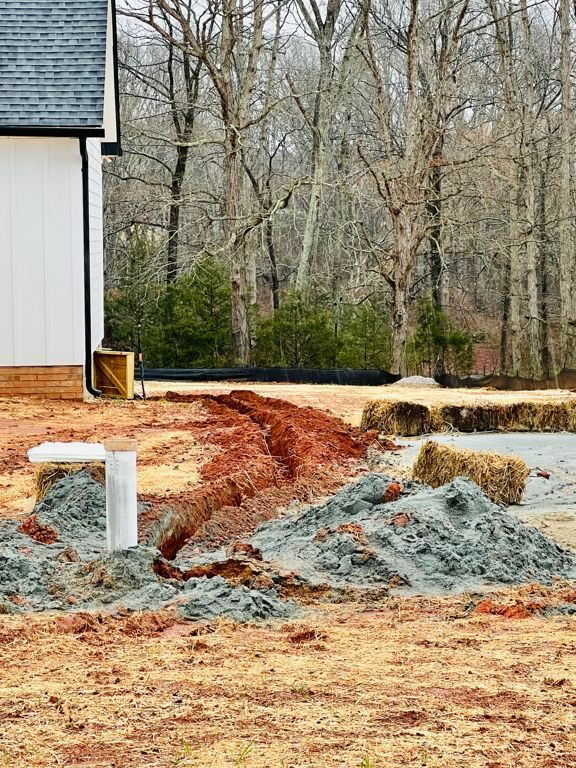Construction materials piled beside a house, with red dirt and a wooded background