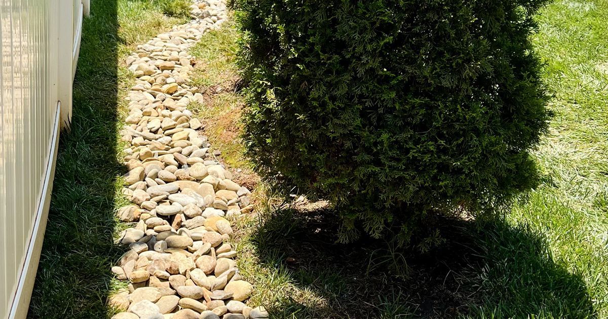 Rock-lined drainage strip beside a white fence and trimmed bushes in a grassy yard