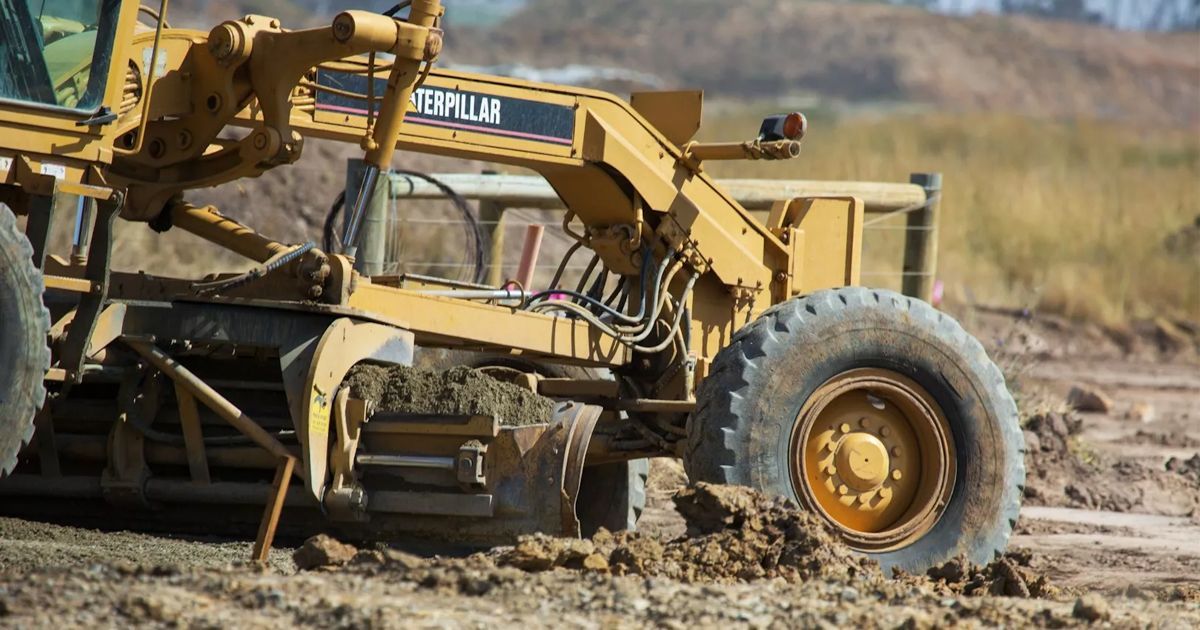 Yellow excavator in muddy field lifting a large tire near construction equipment.