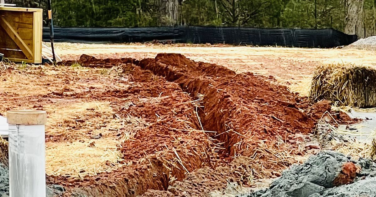 Freshly dug red dirt trench and gravel path at a construction site beside hay bales and fencing