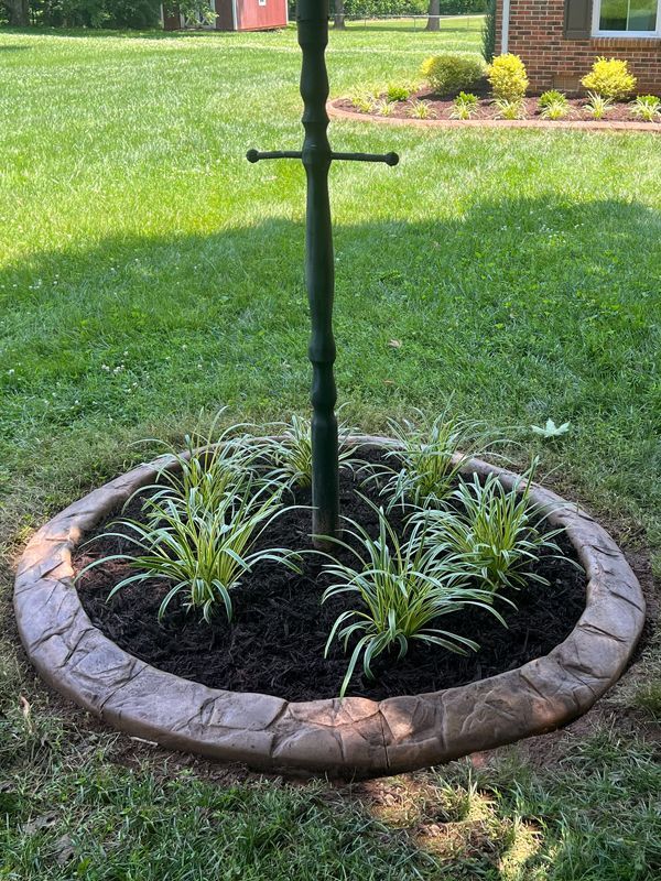 Circular stone planter with black mulch and small green plants around a black pole on a lawn