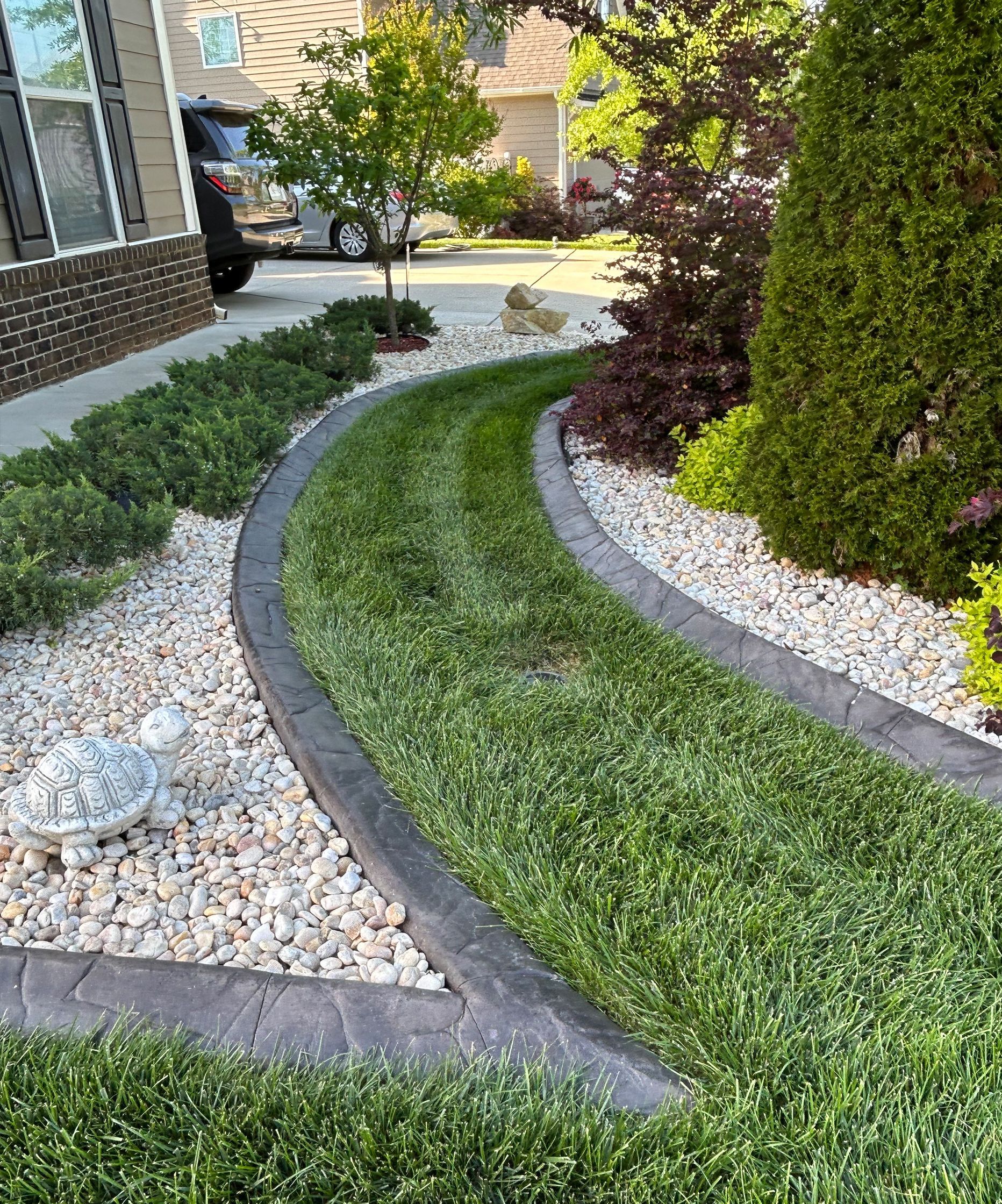 Curved stone-bordered garden beds with white gravel, green grass, and shrubs beside a house