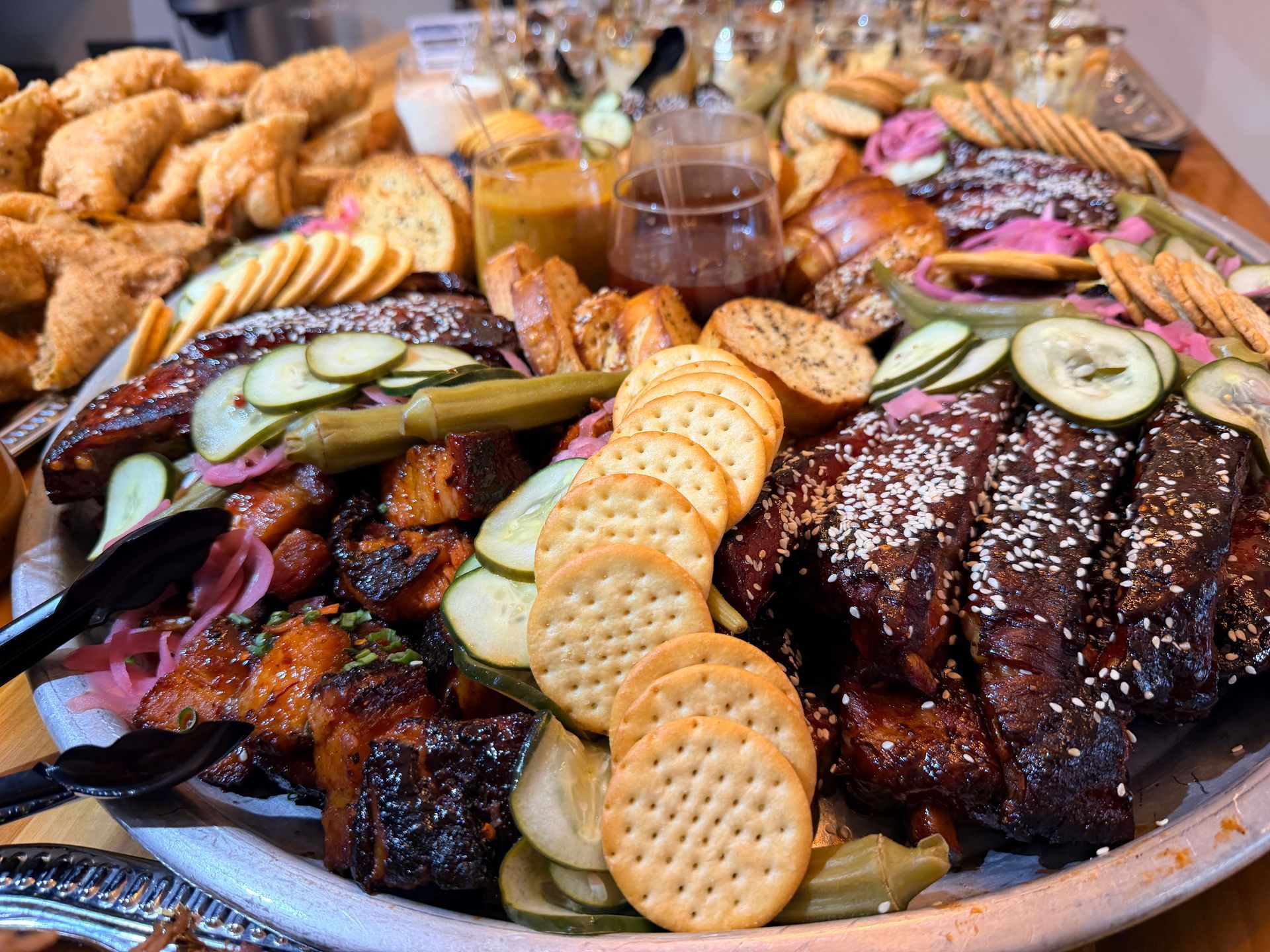 Assorted grilled meats and sides on a buffet table, with sliced cucumbers, crackers, and sesame-topped ribs