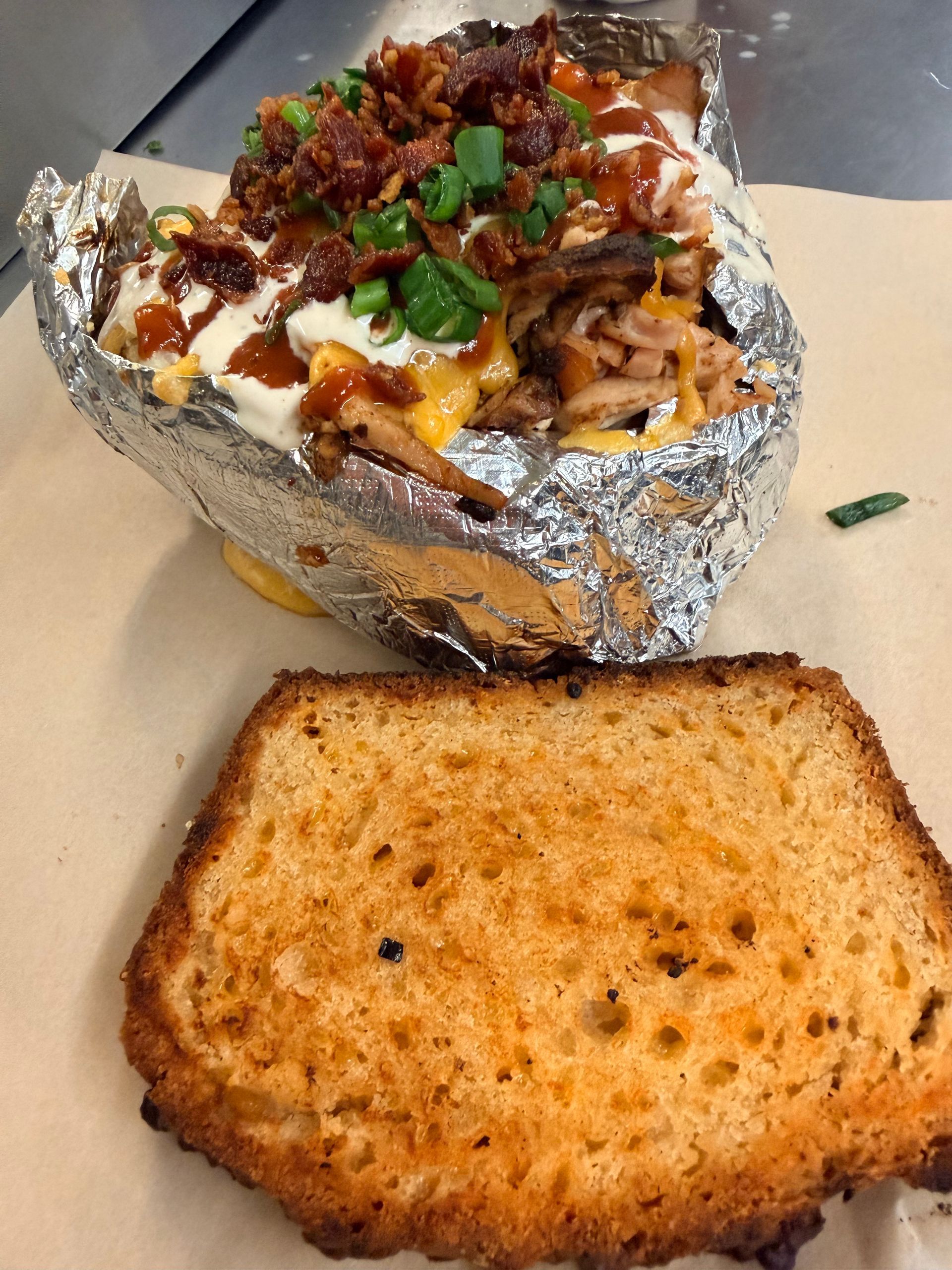 Foil-wrapped loaded baked potato topped with sour cream, bacon, and chives beside a toasted bread slice